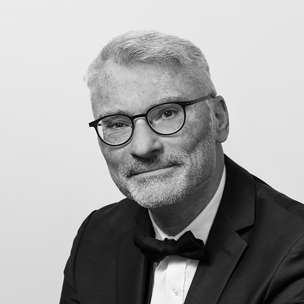 Portrait von Joachim Siegel in schwarz weiß: ein freundlich schauender Mann mit kurzen grauen Haaren, grauem Drei-Tage-Bart und markanter runder, schwarzer Brille im Anzug mit weißem Hemd und Fliege